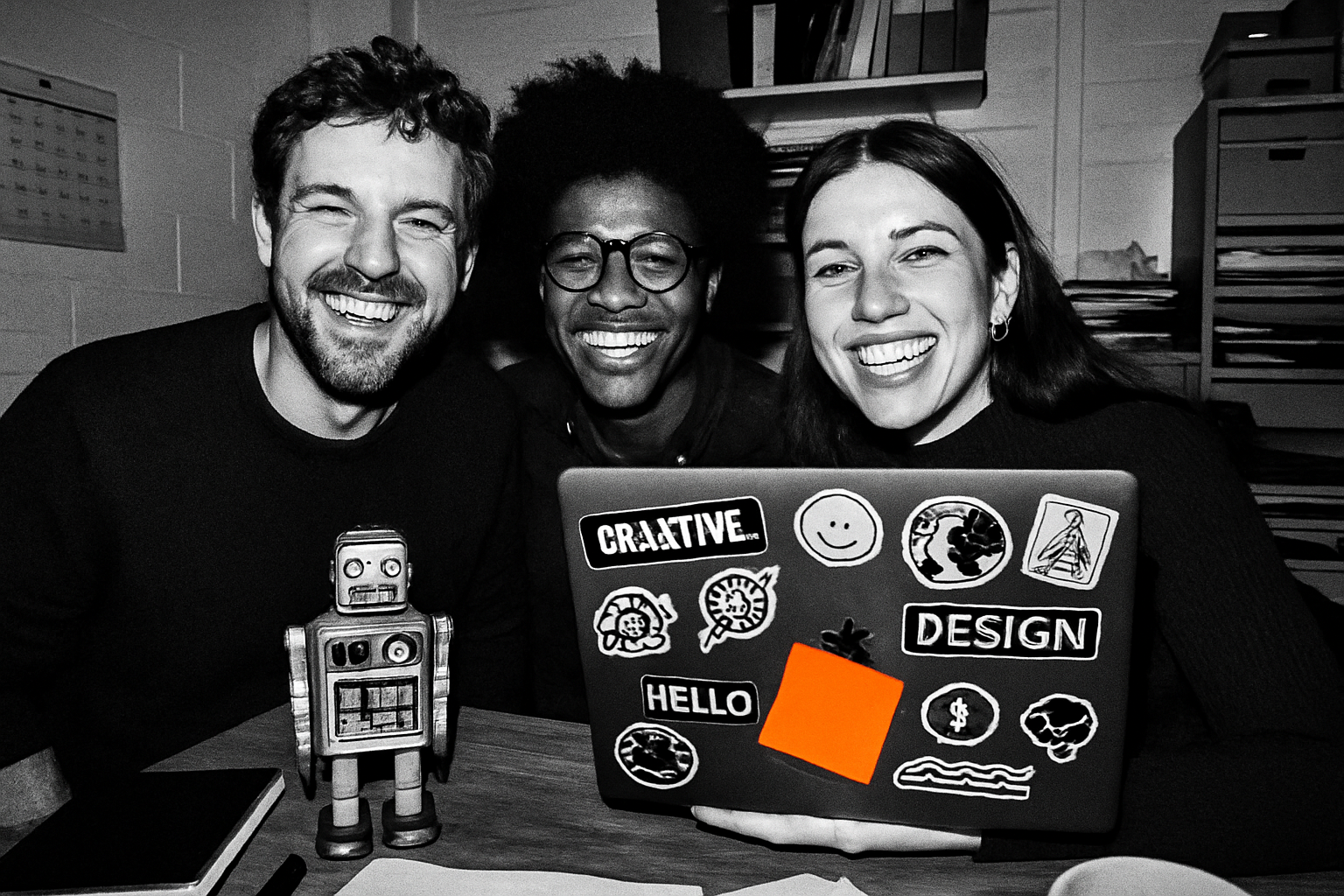 Three grinning agency teammates crammed into a small office with a sticker-covered laptop and a toy robot, shot in harsh black-and-white flash
